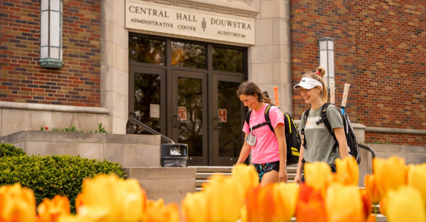 Two students walking in front of Central Hall, with a bed of yellow tulips in the foreground.
