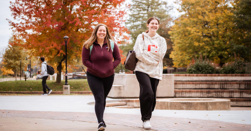 Two students walking across campus framed by fall-colored trees.