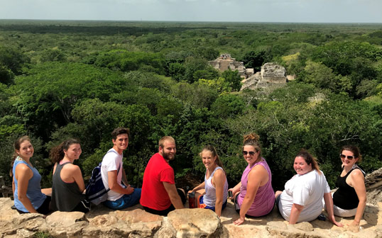 Associate professor of accounting Maggie Schlerman poses for a photo with her class during a trip abroad. In the background are a large forest and pyramids.
